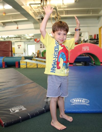 A young boy is enthusiastically holding his hands in the air at a Columbus gym in New York, enjoying the benefits of gymnastics for children.