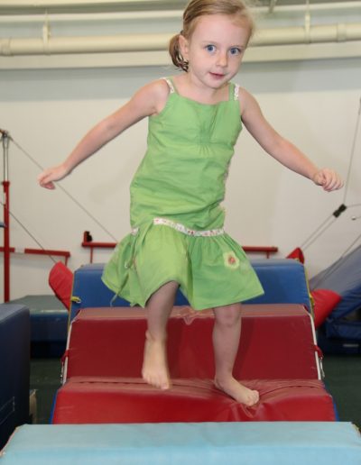 A little girl in a green dress is jumping on a balance beam at a Kid Party.